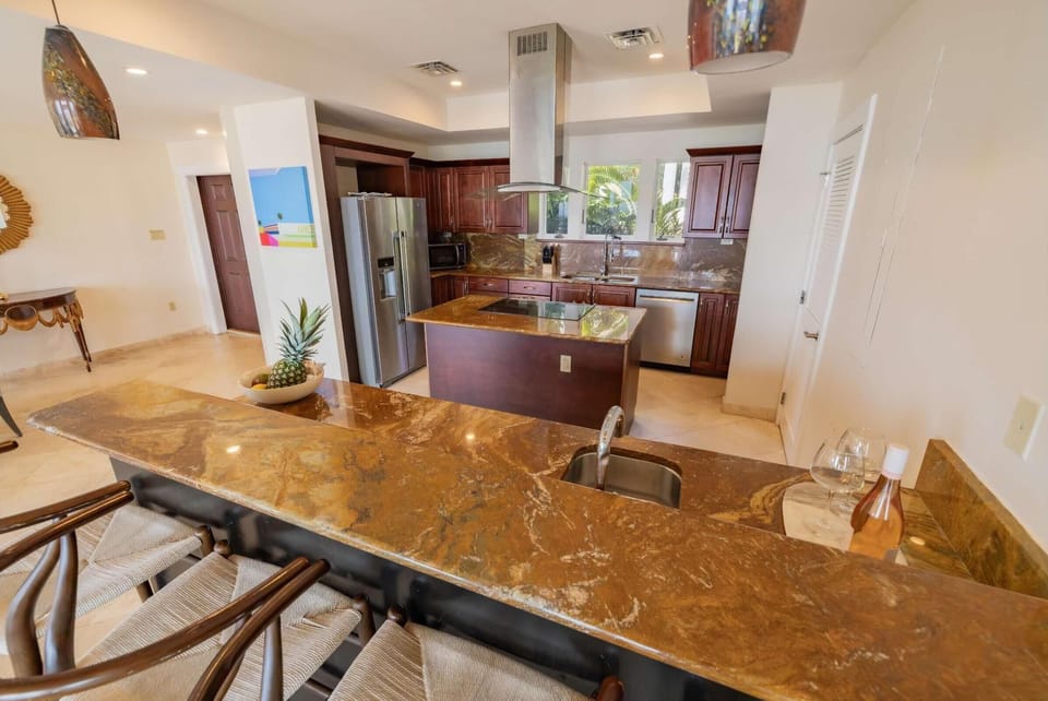 Brown marble breakfast bar with four woven-seat wooden stools overlooks the kitchen island and stainless appliances.