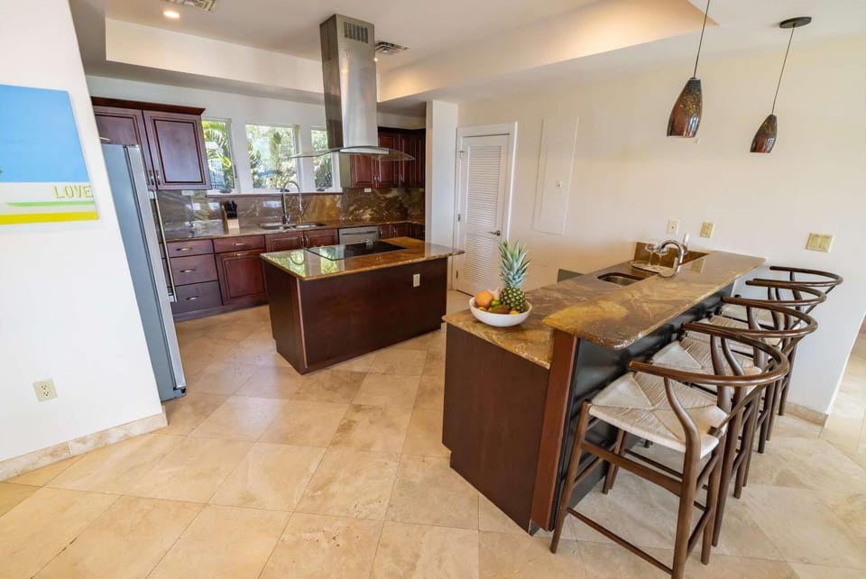 Kitchen island with cooktop and vent hood, dark wood cabinets, and a breakfast bar with four woven-seat stools.