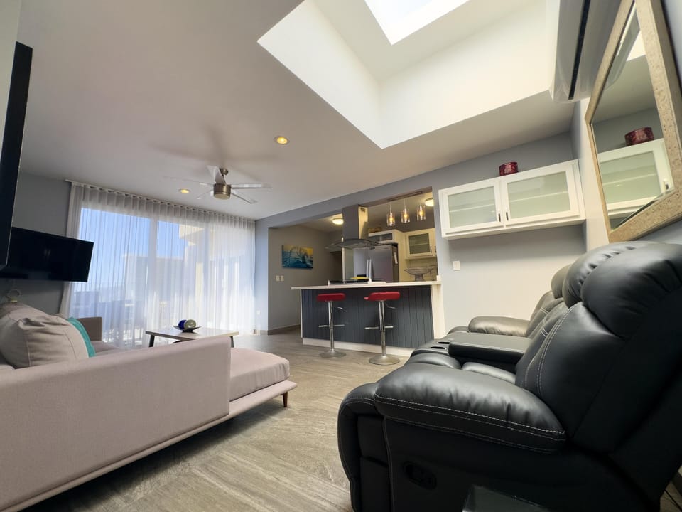 Black leather recliners face a wall-mounted TV opposite a pale pink sofa, beneath a ceiling fan and skylight.