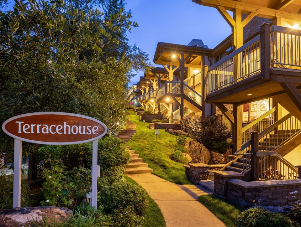 Terracehouse property entrance at dusk, featuring timber-frame architecture with illuminated balconies, landscaped pathways, and welcoming signage nestled among mature trees.