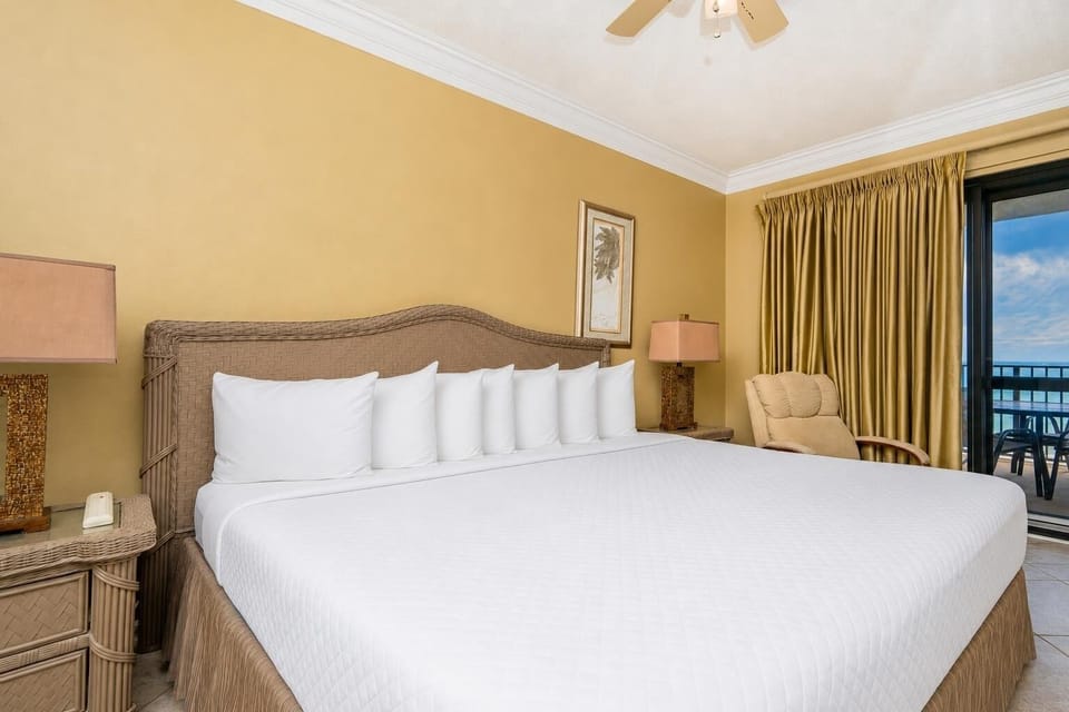 Spacious master bedroom features a king bed with white linens, a cozy armchair, and access to a balcony with gulf views