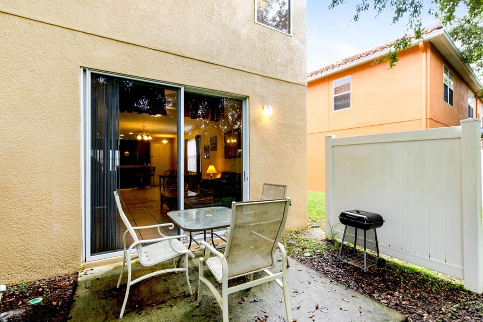 Concrete patio with glass-top table, four mesh chairs, and a compact black charcoal grill by a white privacy fence.