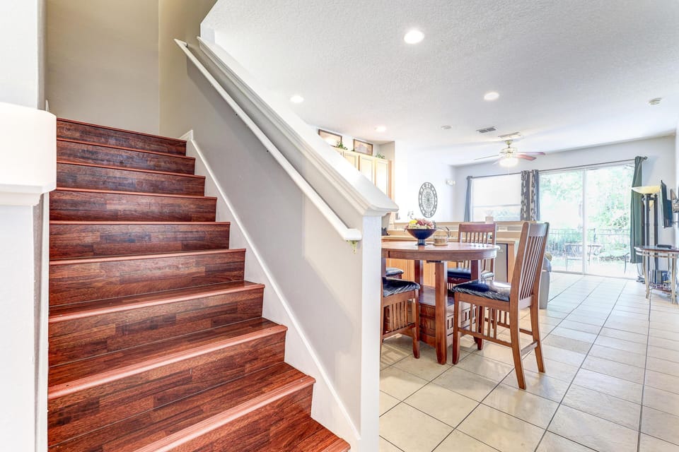 Dark wood stairs lead down to the open living area with tiled floors, a round wooden dining table, and sliding glass doors.