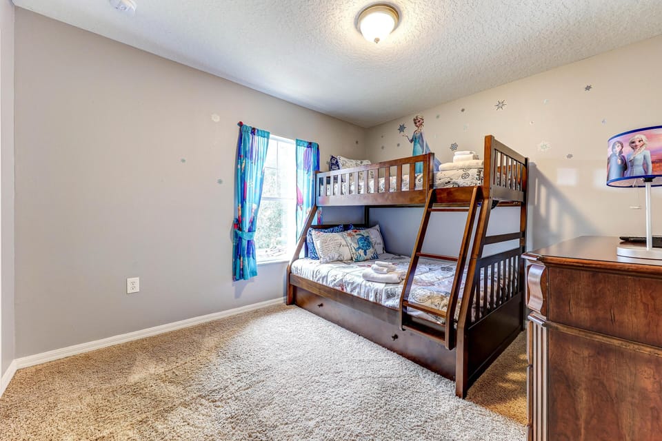 Carpeted bedroom with dark wood bunk bed featuring ladder, patterned bedding, a matching dresser, and a window with blue curtains.