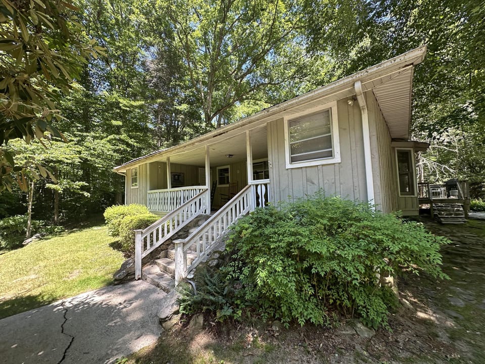 Side view of the cottage with pale green board-and-batten siding, stone steps, and a covered porch shaded by tall trees.