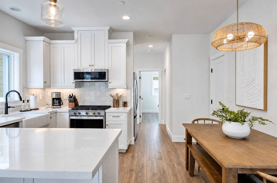 Bright, spacious kitchen and dining area with white cabinetry, modern appliances, and coastal-inspired fixtures. Enjoy preparing meals on gleaming countertops while sunlight fills your open-plan living space.