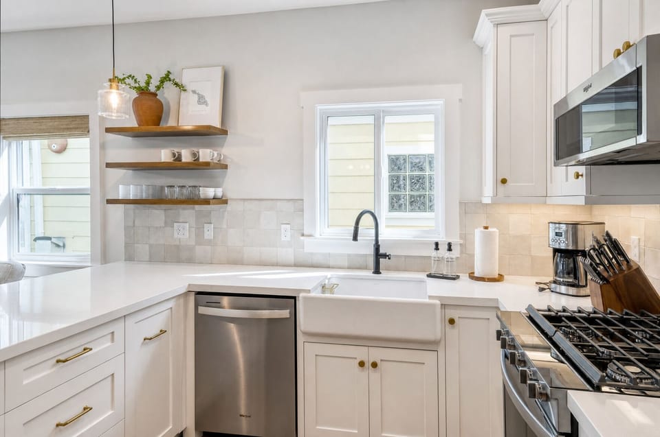 This bright, welcoming kitchen invites you to prepare meals with ease, featuring sleek white cabinetry, stainless appliances, open wood shelving, and a spacious farmhouse sink that makes cooking feel effortless.