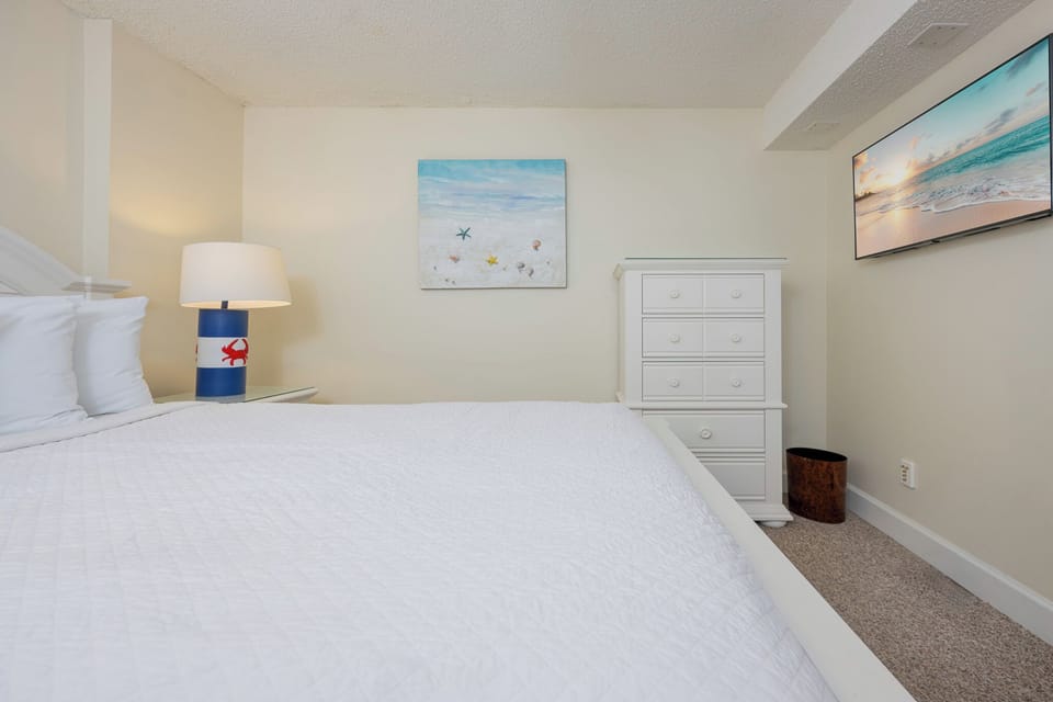 This bedroom features a white quilted bedspread, a mounted TV, and beach-themed artwork for a relaxing atmosphere