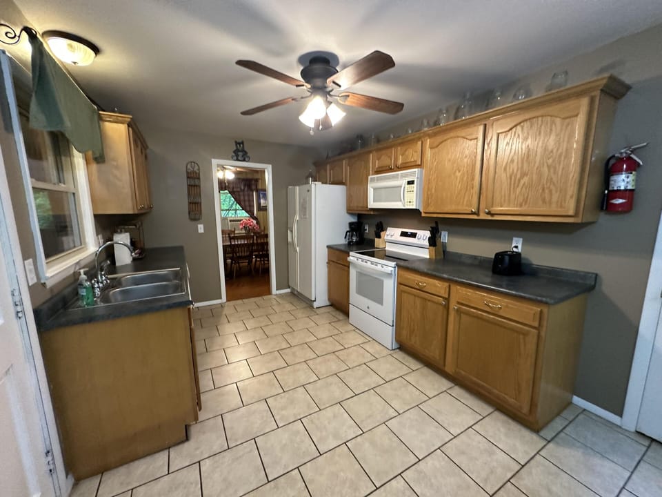 Galley kitchen with oak cabinets, tile flooring, white appliances, and a ceiling fan overhead.