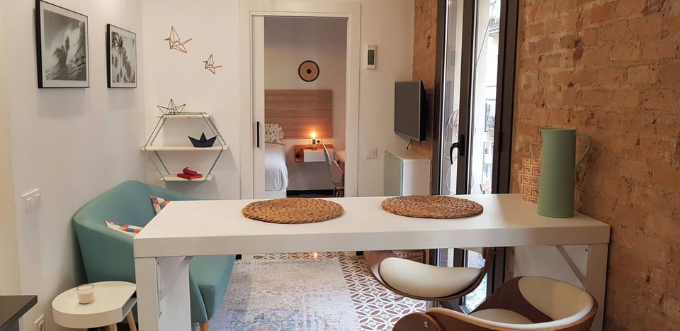 The living space features a white dining table with two chairs. The table has two woven placemats, a light blue sofa with patterned pillows, and a white side table with a lit candle. The dining table is situated in front of a window that looks out onto a 