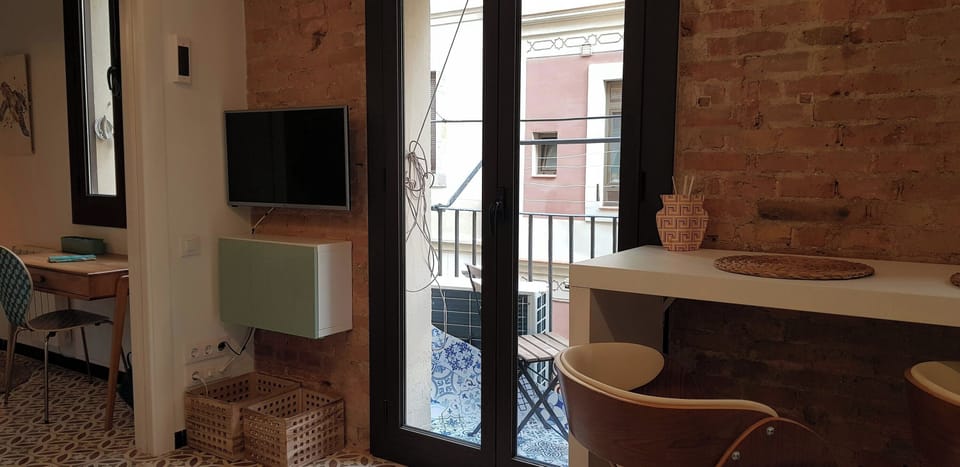 The living space features a flat-screen TV mounted on an exposed brick wall, and a small balcony with a view of the neighboring building, accessible through a large window. The room is decorated with modern furniture and two wooden chairs, and the floors 