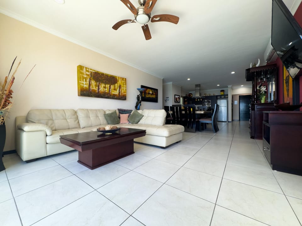 Cream leather sectional and dark wood coffee table sit on glossy white tile, with dining area and kitchen in background.