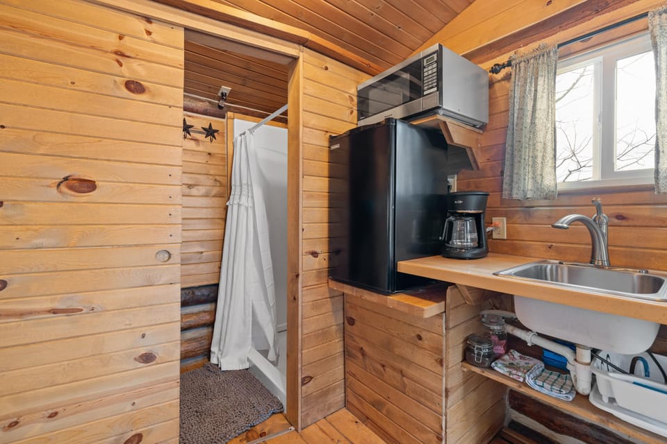Compact kitchenette features a black mini fridge, microwave, coffee maker, and a stainless steel sink beneath patterned curtains.