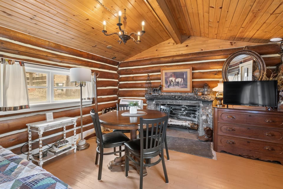 Dining area with round wood table, four black chairs, stone fireplace, and wooden log cabin walls.