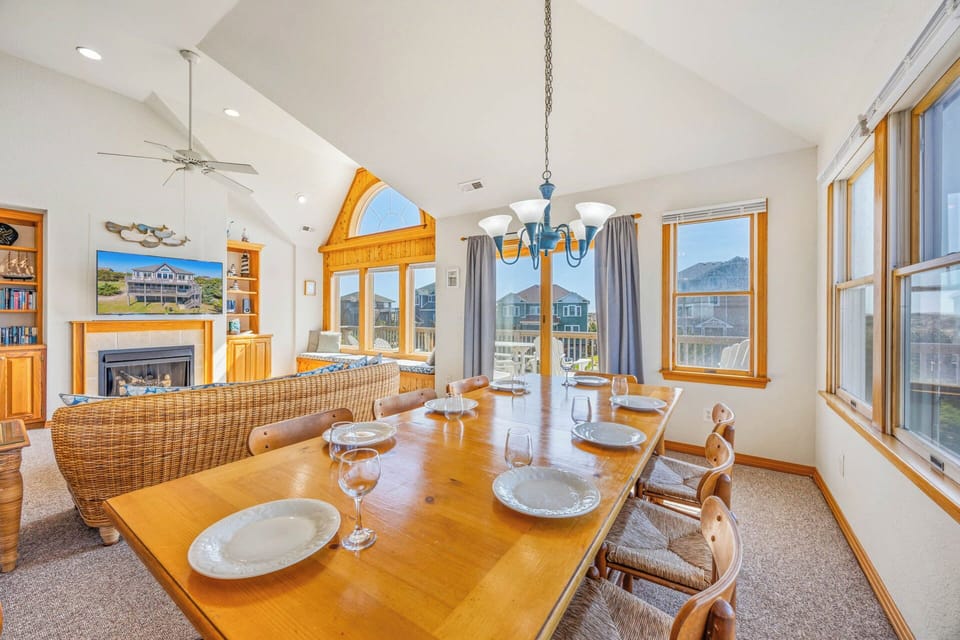 Gather around the warm, inviting dining table where meals become memories. The open-plan living space, cathedral ceilings, and natural light create a welcoming atmosphere perfect for your vacation gatherings.