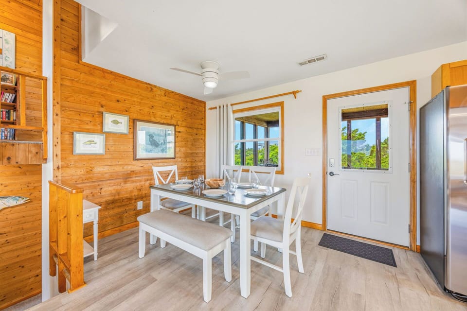 Gather around the cozy dining table where warm wood tones meet coastal charm. Your meals here are framed by natural light streaming through cottage-style windows, inviting you to savor every moment together.