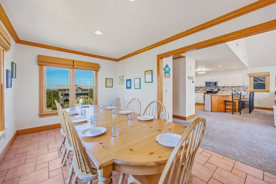 Gather around this inviting dining table where sunlight streams through windows framing coastal views. The open layout flows into your fully-equipped kitchen, perfect for preparing family meals and creating vacation memories together.