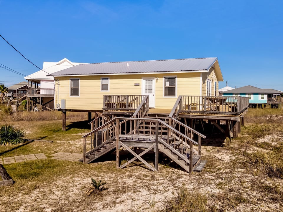Charming yellow beach house elevated on stilts with wooden deck and stairs, surrounded by coastal vegetation under clear blue skies.