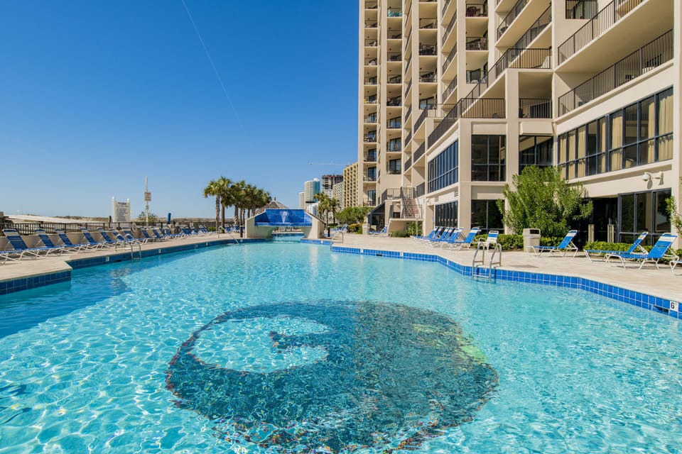 Outdoor Pool, Beach Chairs, Plants, Palm Trees, Phoenix Logo, Clear Water, Sky.