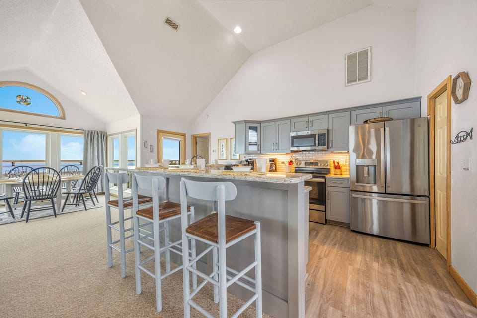 Bright coastal kitchen with vaulted ceilings and ocean views. Granite countertops, stainless steel appliances, and island seating create the perfect gathering space with beach-inspired charm and natural light.