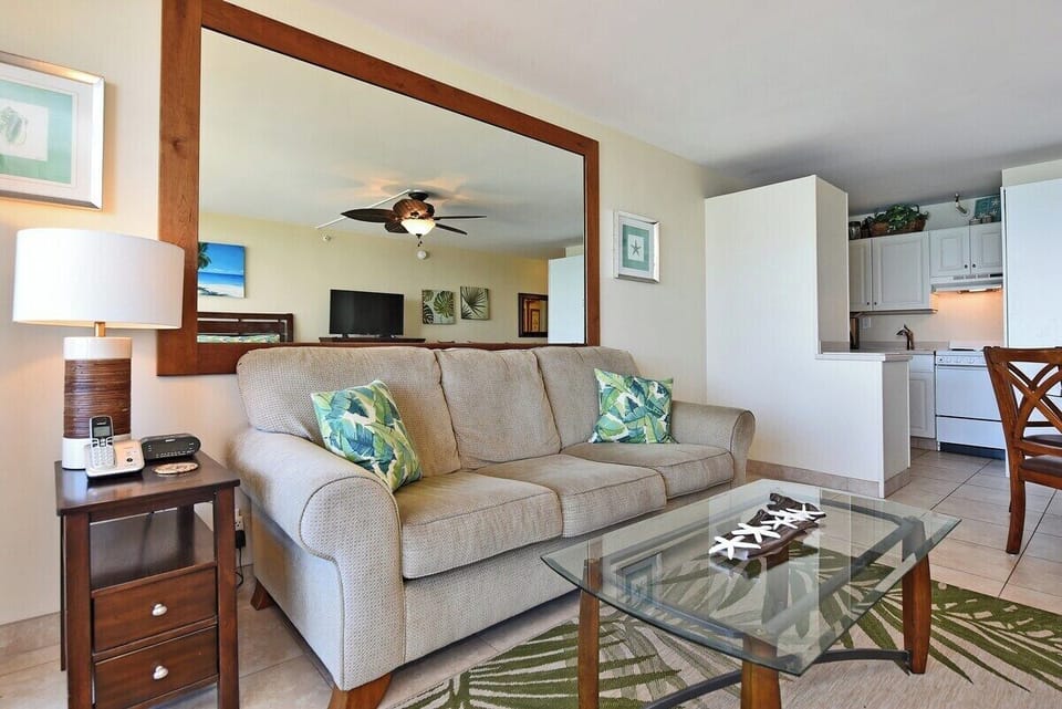 Living room with beige sofa, glass coffee table, and tropical décor, ceiling fan above and open layout leading into the kitchen.