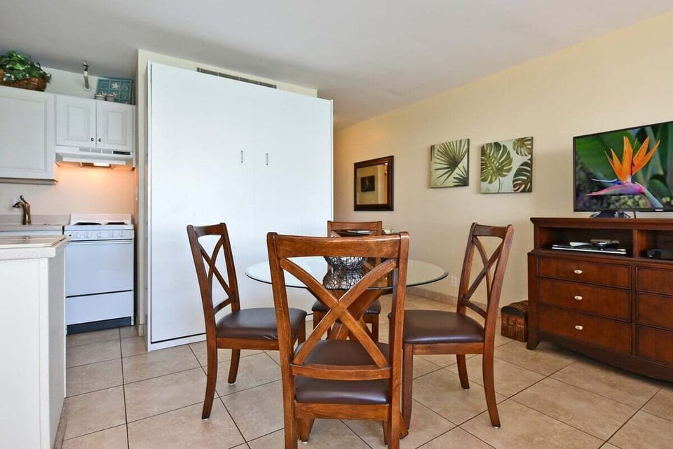 Kitchen and dining space with white cabinetry and appliances, wooden dining table with chairs, dresser with television, and open layout connecting to the rest of the suite.