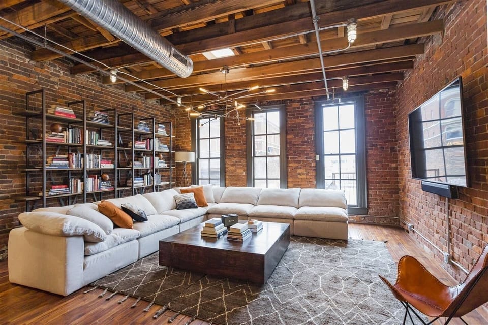 - Industrial loft living room with exposed brick and warm lighting   - Large sectional sofa and tall windows create a relaxing lounge vibe   - Perfect spot for groups wanting a stylish downtown stay