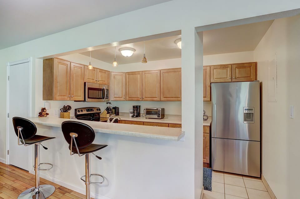Modern kitchen with light wood cabinetry, stainless steel appliances, and a breakfast bar with two black barstools. The room features pendant lighting and a hardwood floor.
