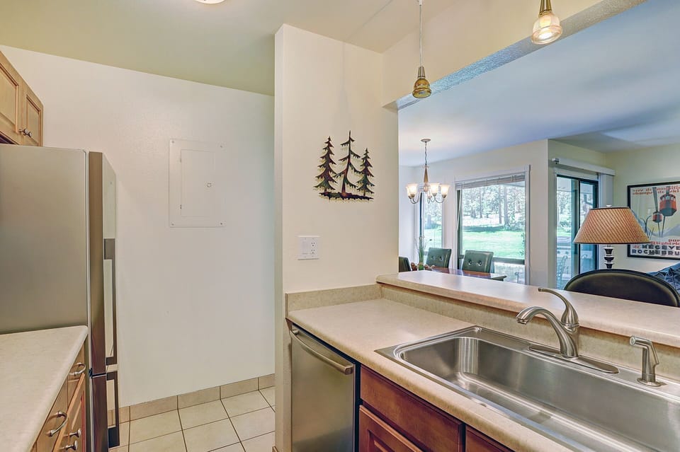 A kitchen area with a stainless steel sink, wooden cabinets, tiled floor, and minimalistic wall decorations, including tree-shaped art. The space opens to a dining area with a view of a grassy outdoor area.