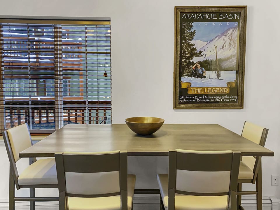 A dining area featuring a wooden table with a brass bowl in the center, surrounded by four chairs. A window with blinds and a framed 'Arapahoe Basin' poster are visible in the background.
