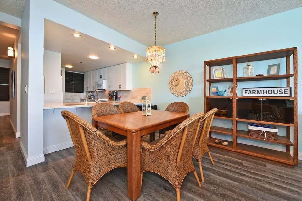 Dining area with wooden table and wicker chairs, light fixture overhead, kitchen with white cabinetry beyond.