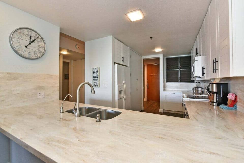 Kitchen countertop with double sink and white cabinets, wall clock and appliances visible down the hall.