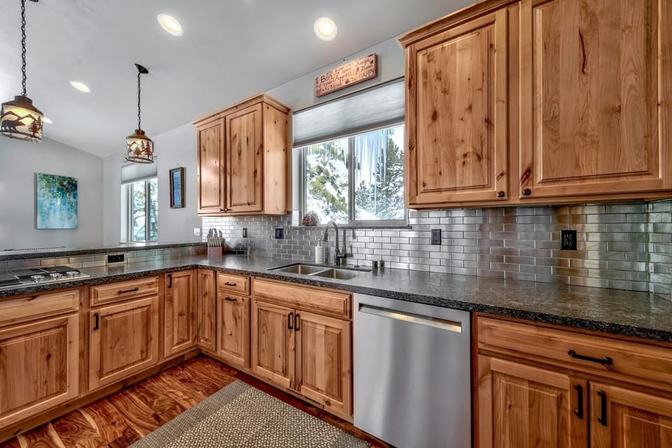 Knotty pine cabinets and stainless steel dishwasher surround a double sink under a window with snowy forest views.