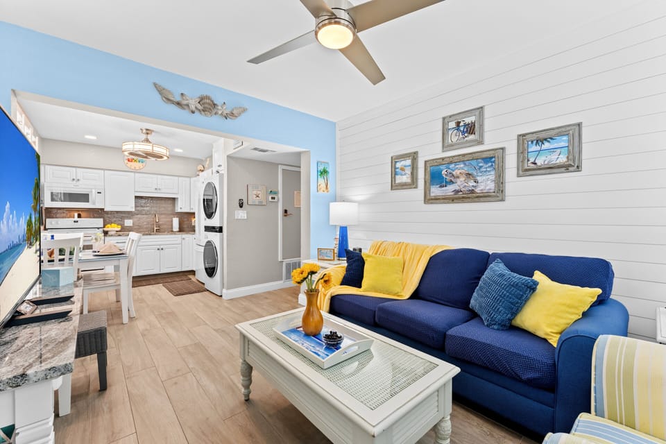 Relax in this cheerful living room featuring a plush sofa, cozy armchair, and smart TV — all wrapped in soft coastal blues and sunny yellow accents.