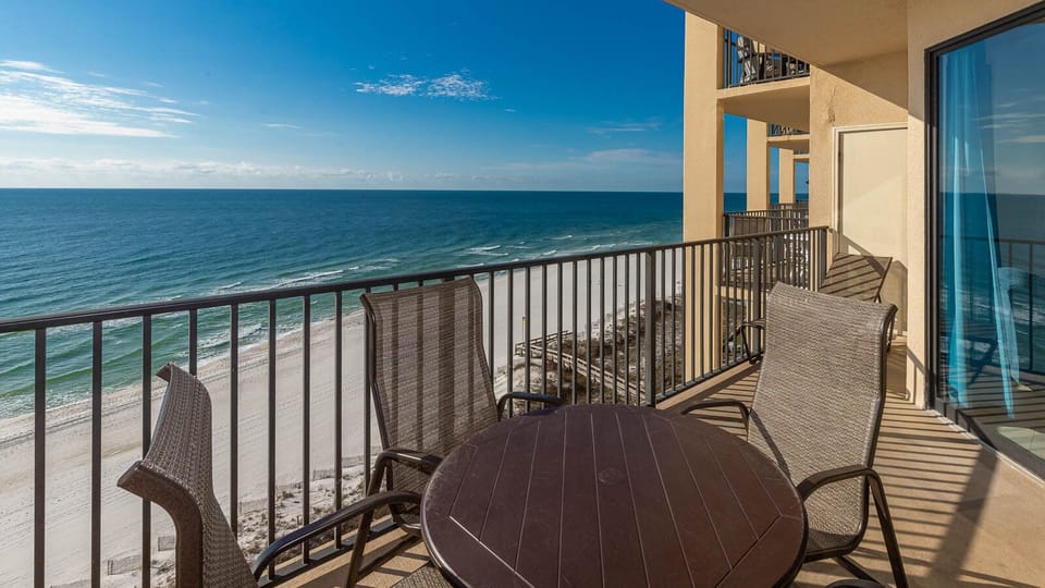 The balcony features a round table and wicker chairs, offering a serene view of the beach and turquoise waters
