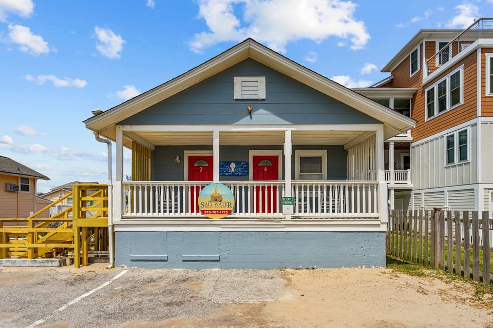 Charming beachfront cottage featuring red doors and covered porch, nestled among coastal homes with convenient parking.
