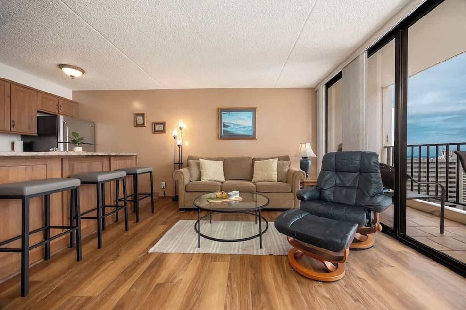 This inviting living space features a pull-out sleeper sofa and a relaxing recliner, a modern kitchen bar with stools for dining, and large glass doors leading to a private balcony with scenic views.