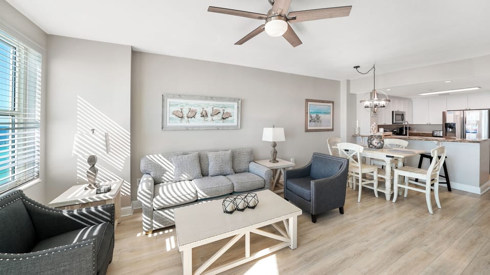 Relax in your bright, coastal-inspired living space where sunlight streams through blinds, creating beautiful patterns across comfortable seating and open kitchen.