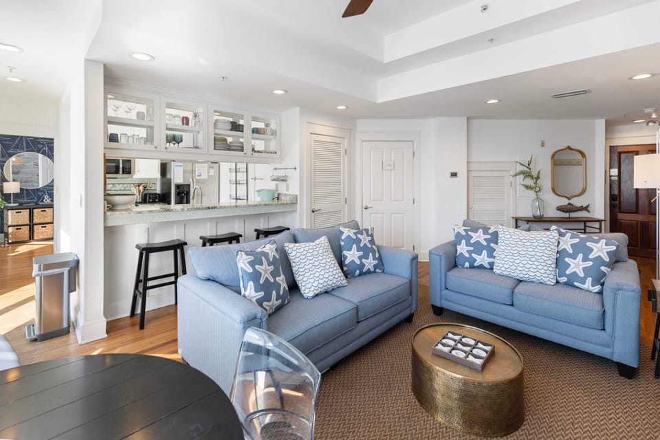 Your bright coastal living space features comfortable blue sofas and a fully-equipped kitchen, perfect for relaxing after beach adventures.