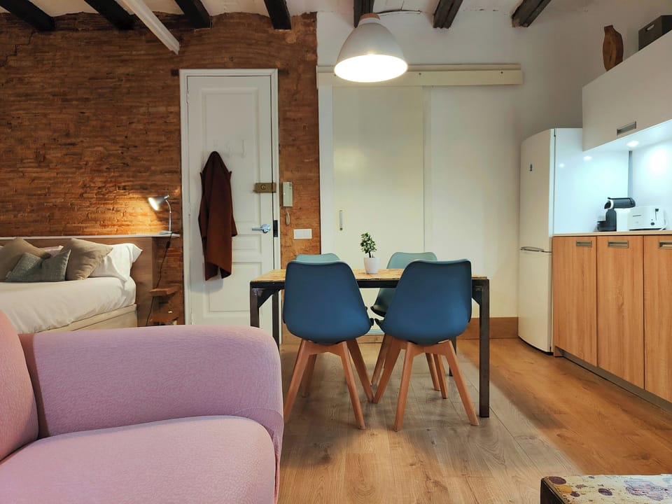 The loft features a light and airy dining area with a wooden table and four blue chairs. The exposed brick wall adds character to the space, while the wooden floor creates a warm and welcoming ambiance. The kitchen, visible in the background, is equipped 
