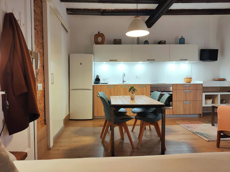 The open-plan living space features a dining table with four chairs positioned in front of a modern kitchen. The kitchen boasts sleek white cabinets, a full-size refrigerator, and a stovetop with an overhead range hood.  Natural light streams in from a wi