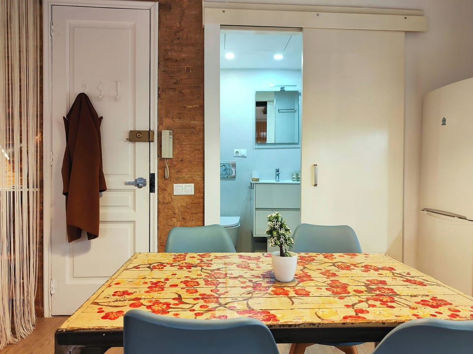 The dining area features a unique, rustic wooden table with a hand-painted floral design, adding a touch of whimsy and charm to the space. Four blue chairs surround the table, perfect for enjoying meals or working remotely. A glimpse into the bathroom thr
