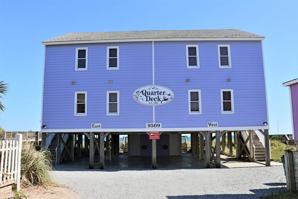 Bright purple beach house elevated on stilts with Quarter Deck signage, featuring East and West units for your coastal getaway.