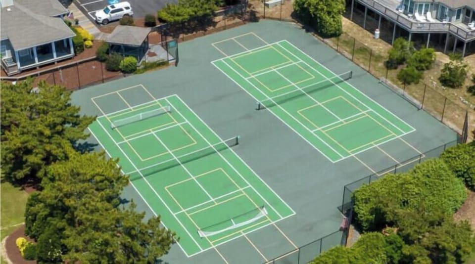 Aerial view of the community's tennis and pickleball courts, surrounded by mature trees and neighboring vacation homes in the residential area.