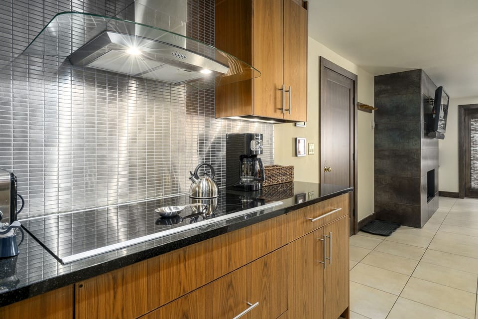 Modern kitchen with a stainless steel stove, glass range hood, and wooden cabinetry. A coffee maker and kettle are placed on the black granite countertop. Tile flooring and a wall-mounted TV are visible.