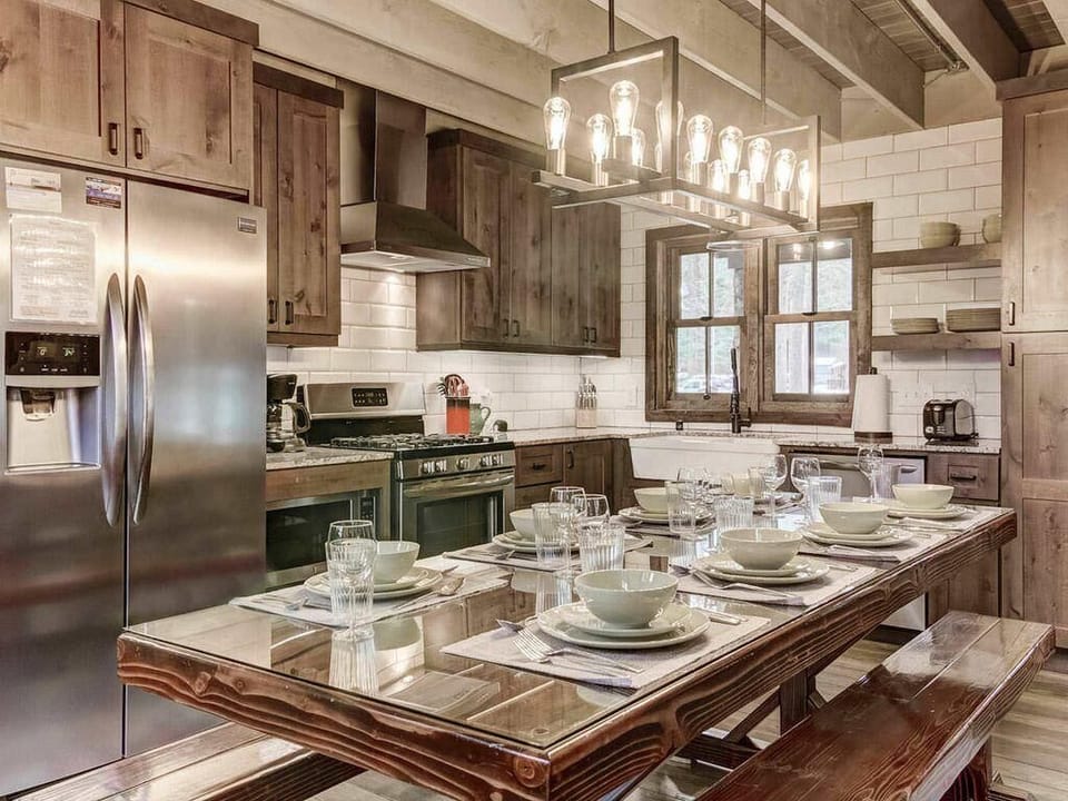 Glass-covered wooden dining table set for eight beneath industrial-style Edison bulb chandelier in kitchen.