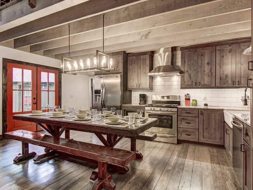 Dark wood cabinetry and stainless appliances line the kitchen, with a long wooden dining table set beneath exposed beams.