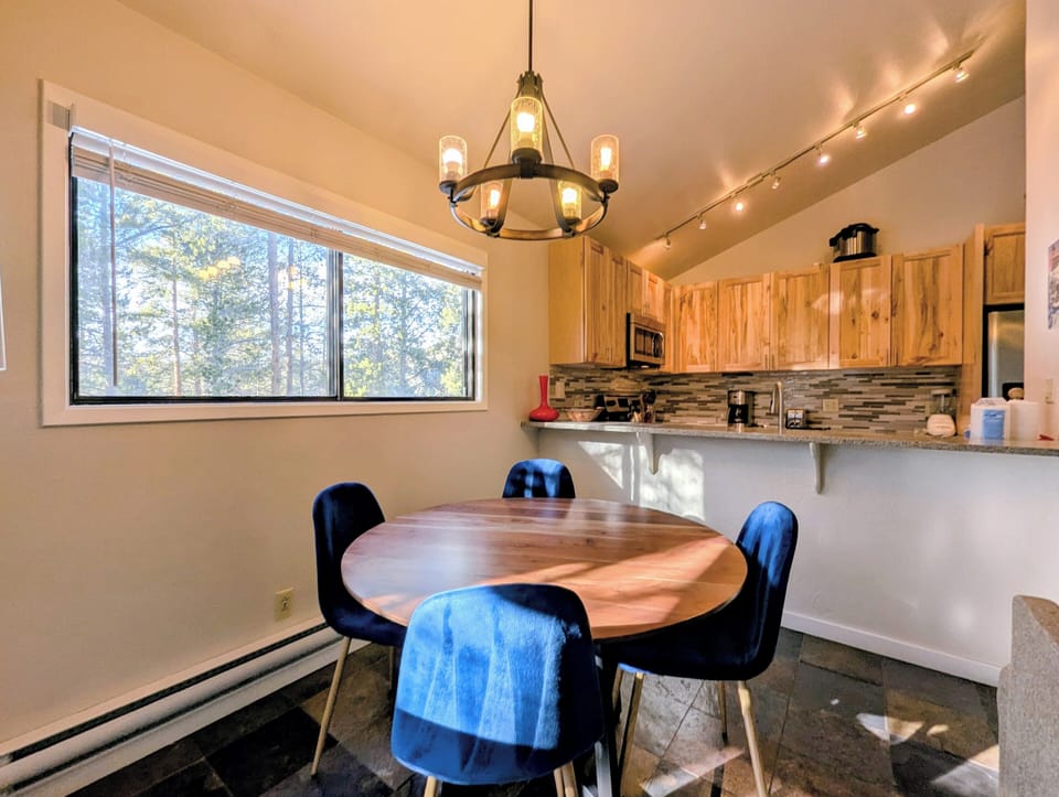 Gather around your warm wood dining table in this bright kitchen, where natural light streams through large windows revealing peaceful forest views.
