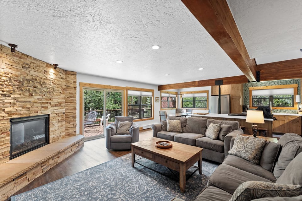 Sink into plush sectional seating by your stone fireplace, where warm wood beams frame forest views through expansive windows.