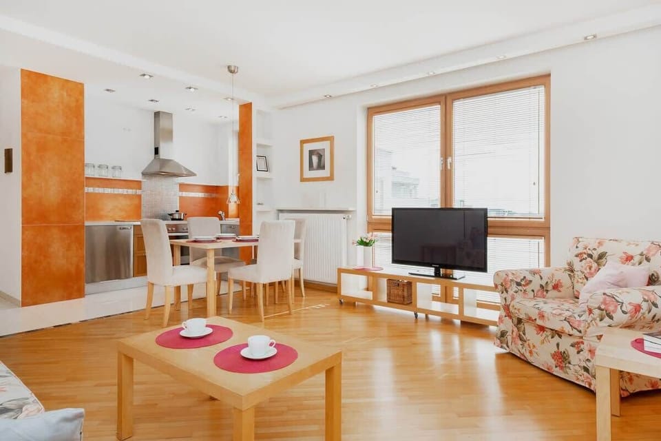 A spacious living room with a dining area, featuring wooden flooring, a cozy sofa bed, and a dining table for four. The large windows let in plenty of natural light.